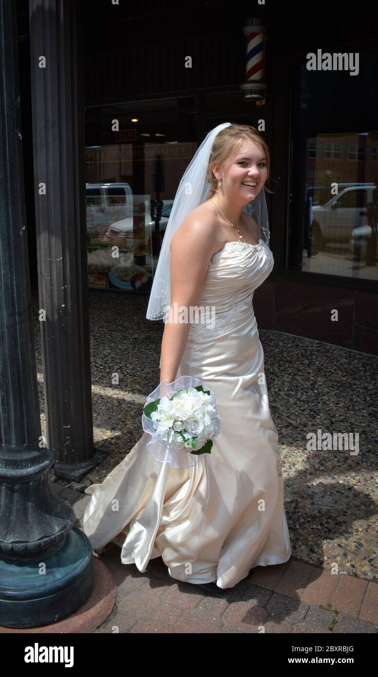 A young Caucasian bride in white strapless wedding gown with veil, smiles holding bouquet while walking solo down a sidewalk in downtown St. Cloud, MN Stock Photo