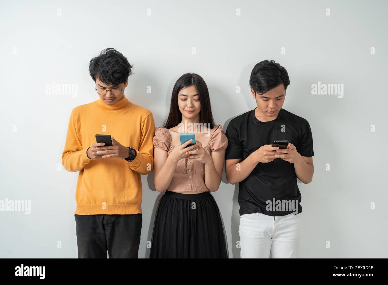 Three young Asian people use their smart phones while standing against ...
