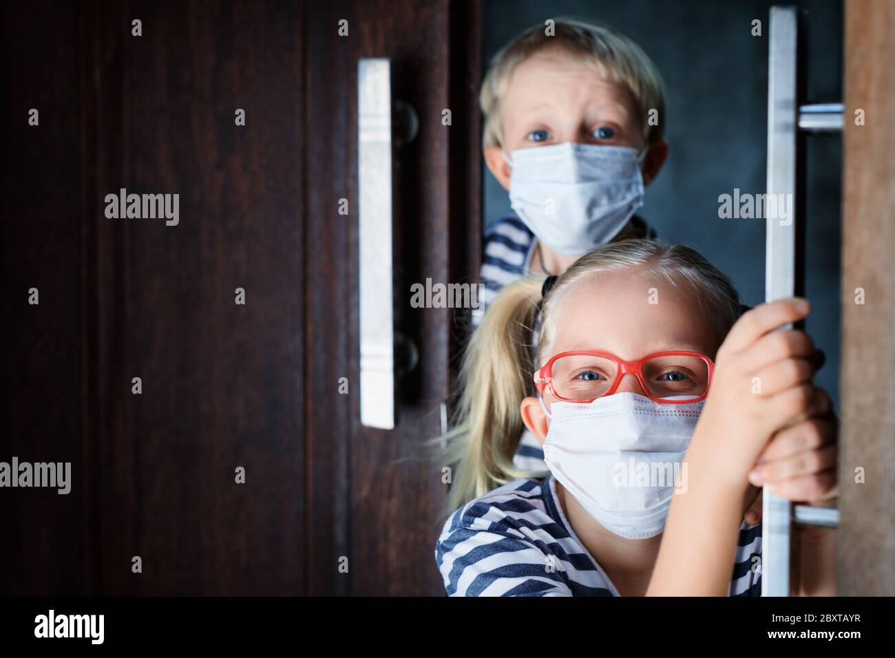 Little children looking out opened door after staying at home due banned street activity. Kids wearing medical face masks go out for outside walk. Stock Photo