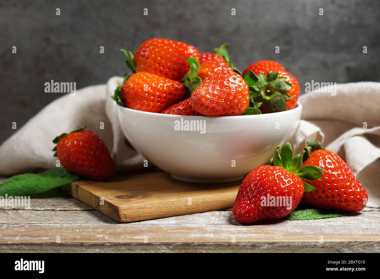 Strawberry concept with a group of ripe red strawberries in a white bowl close up frontal view on a vintage rustic wooden table and grey background. Stock Photo