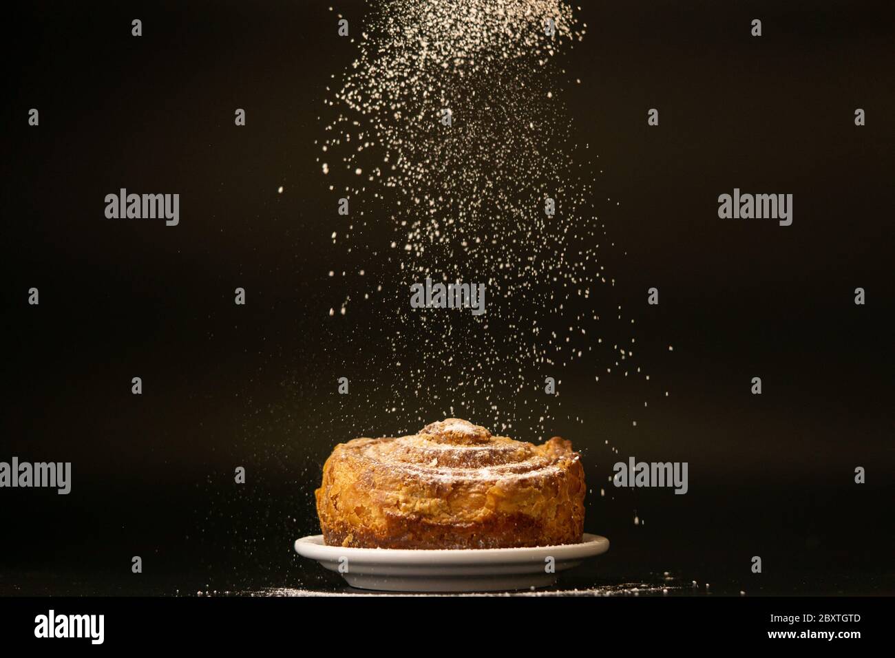 Icing sugar is sprinkling over fresh bundt cake. Icing sugar falls down, freezing motion. Copy space, black background. Bakery concept. Sweet bread on a white plate on a black background. Stock Photo