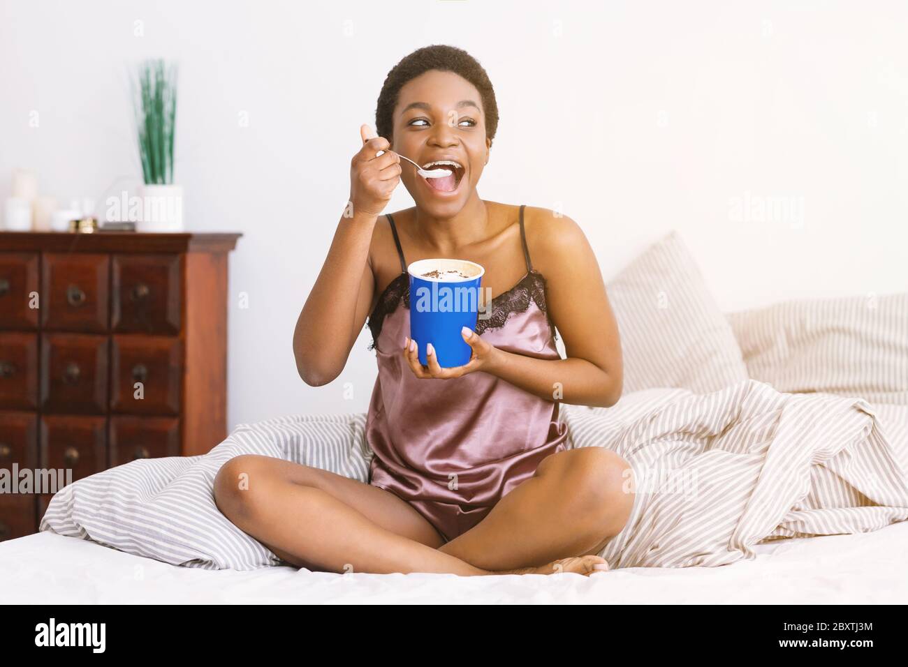 Fun and sweets. African american girl in pajamas eating ice cream from bucket on bed Stock Photo