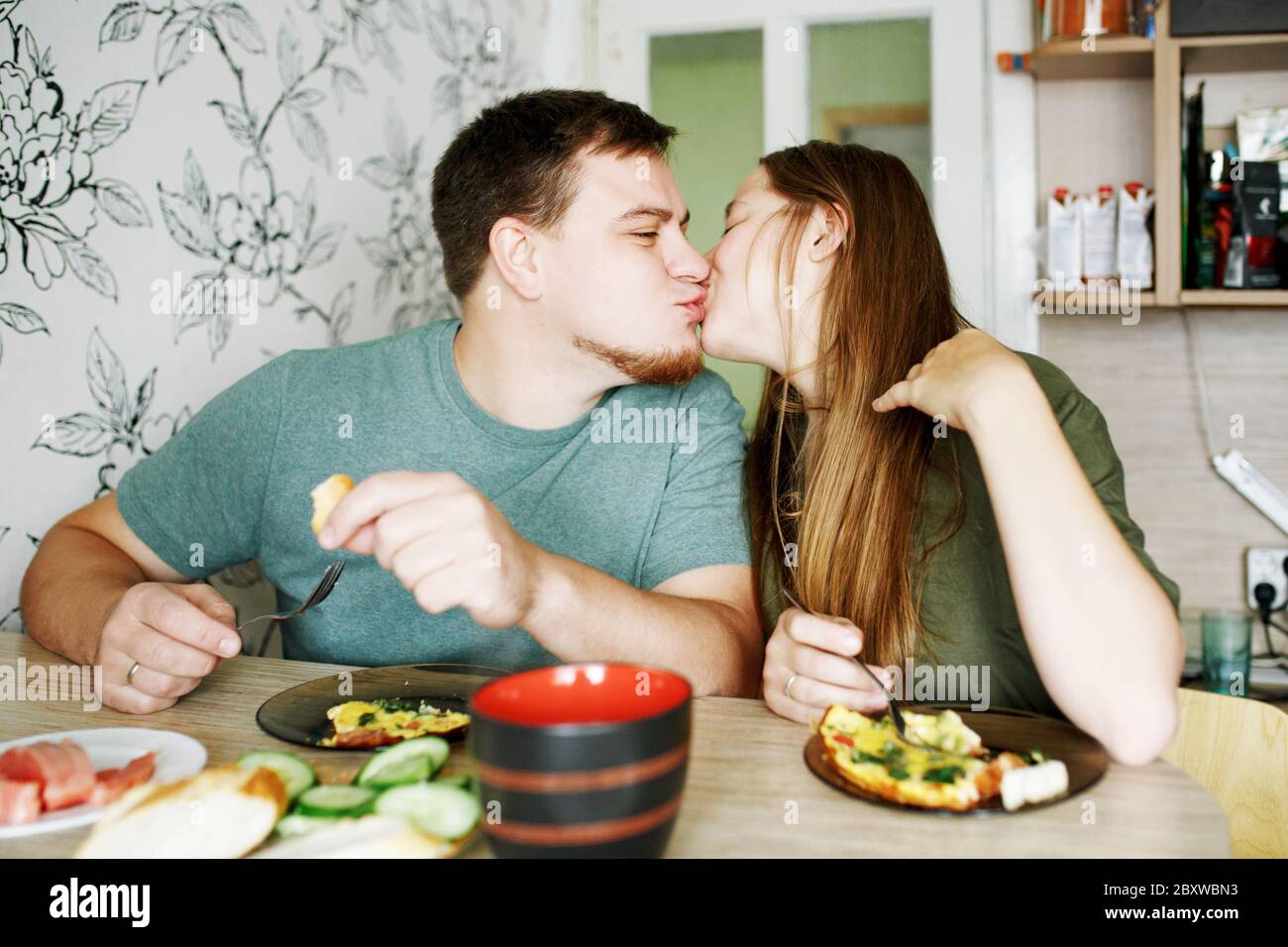 Young candid family eating omelette eggs for breakfast at home in apartment, happy girlfriend and boyfriend kissing Stock Photo