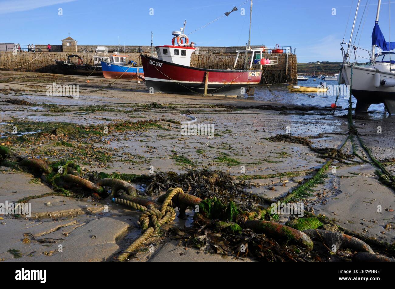 Rusty Chain and seaweed on beach,  mooring fishing boats and yachts in harbour with the old quay in the background, at Hugh Town, St Mary's one of the Stock Photo