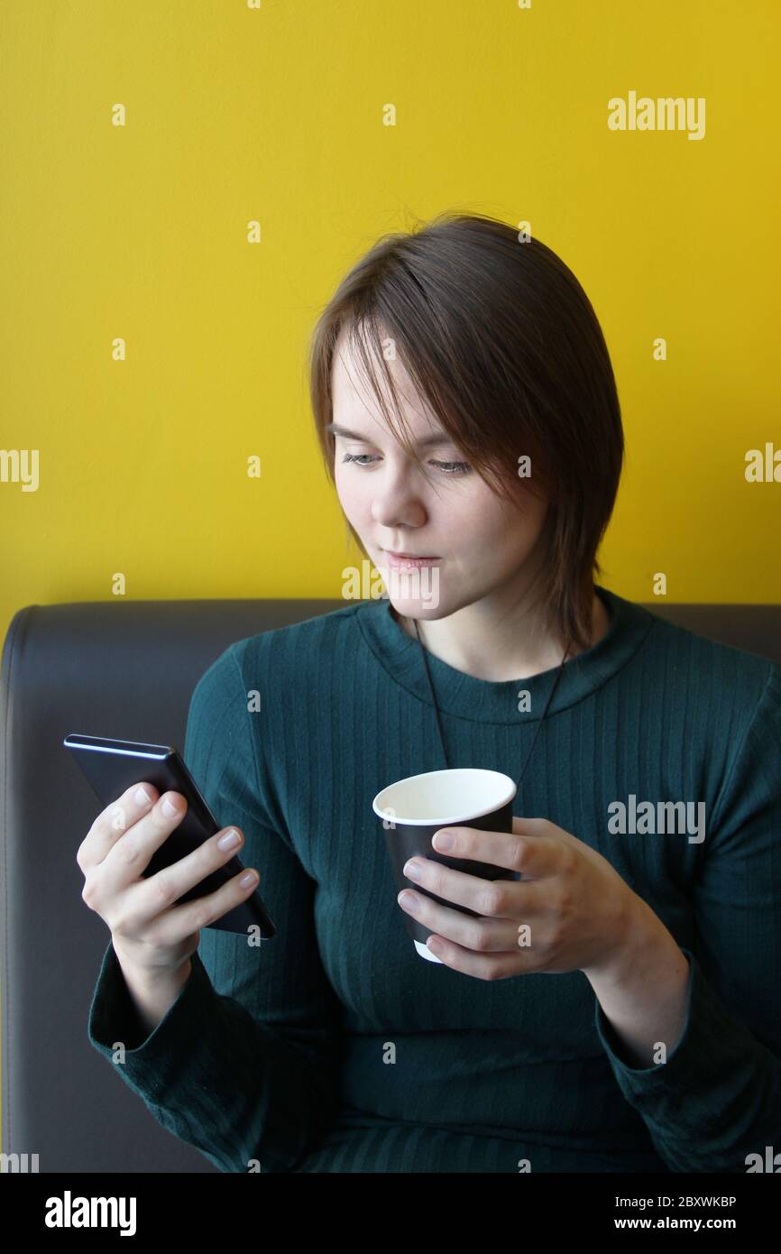 A young beautiful girl of European appearance in a blue jacket sits in a cafe with a mobile phone on the couch against the background of a yellow wall. Holding a paper cup with coffee. Stock Photo