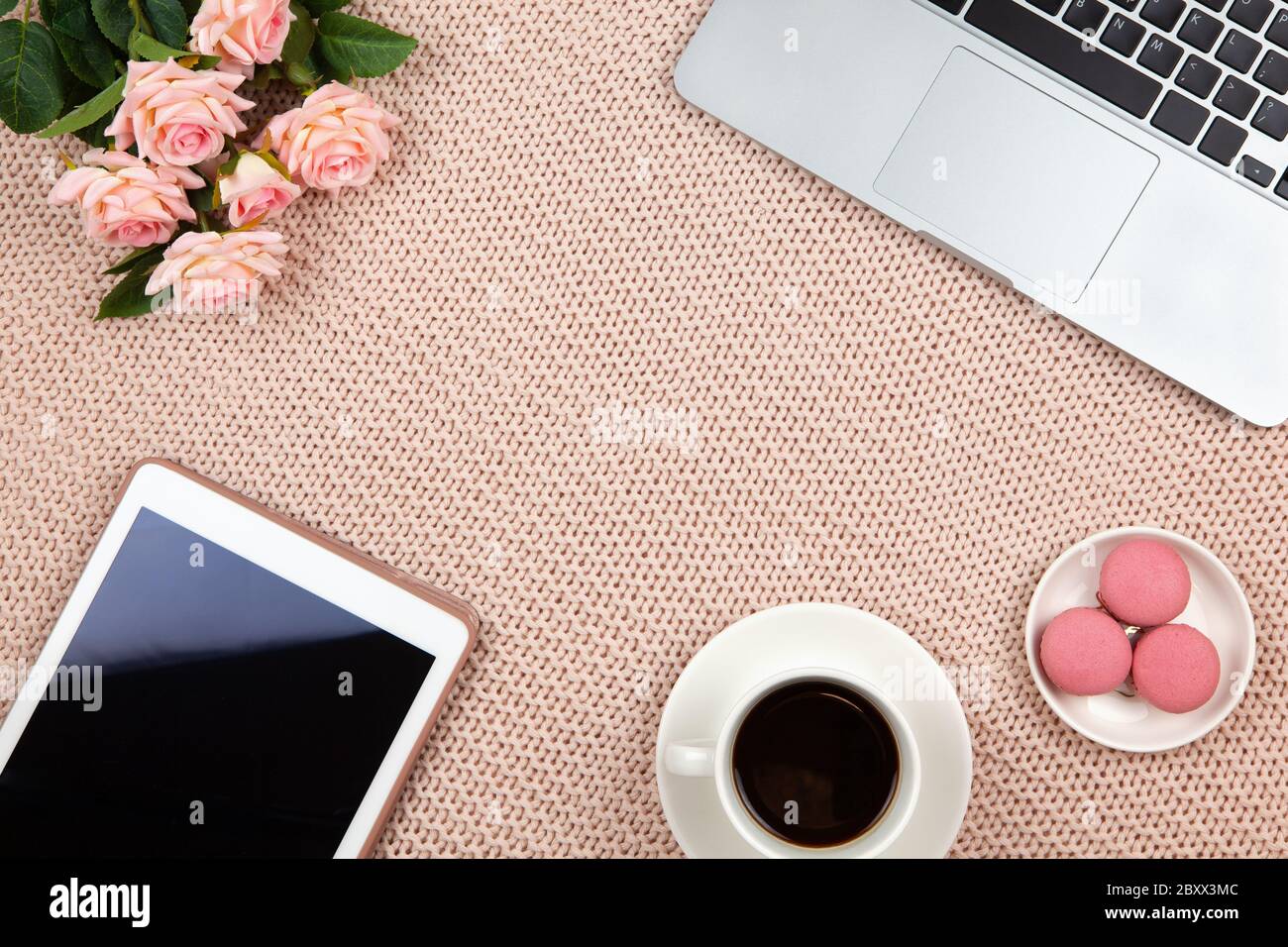 Work from home concept. Modern female working space, top view. Laptop, coffee, cakes, roses, tablet on knitted blanket, copy space, flat lay. Desktop Stock Photo