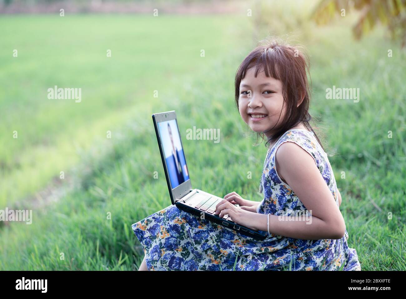Happy little child girl learning outdoor by studying online and working on laptop in green field Stock Photo
