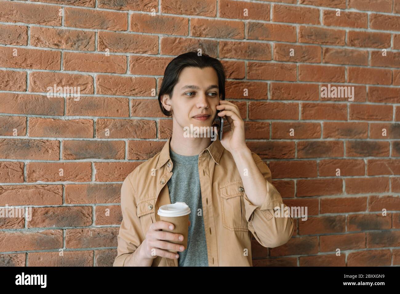 Man using mobile phone, holding cup of coffee in cafe. Hipster guy wearing stylish casual clothing holding smartphone, talking near wall. Coffee break Stock Photo