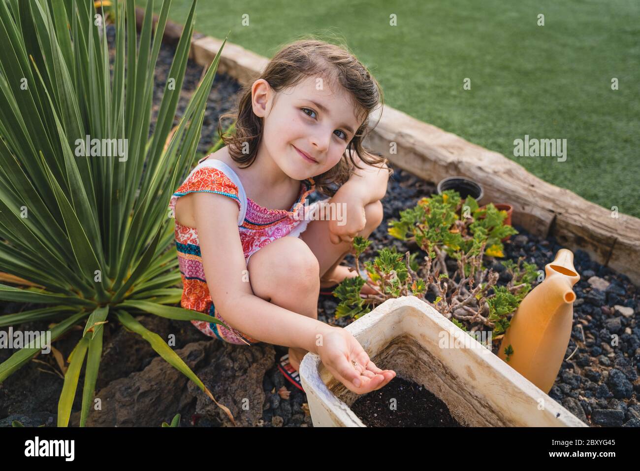 cute little girl in backyard garden holds plant seeds in her hand into pot, smiling to camera sitting down on the ground Stock Photo