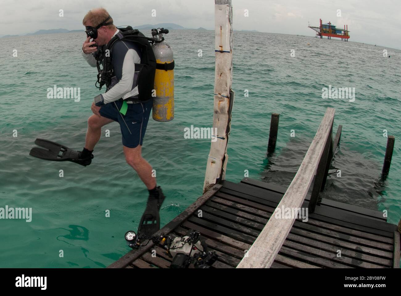 Man in scuba gear jumping into water from jetty with oil rig in background, Mabul Island, Sabah