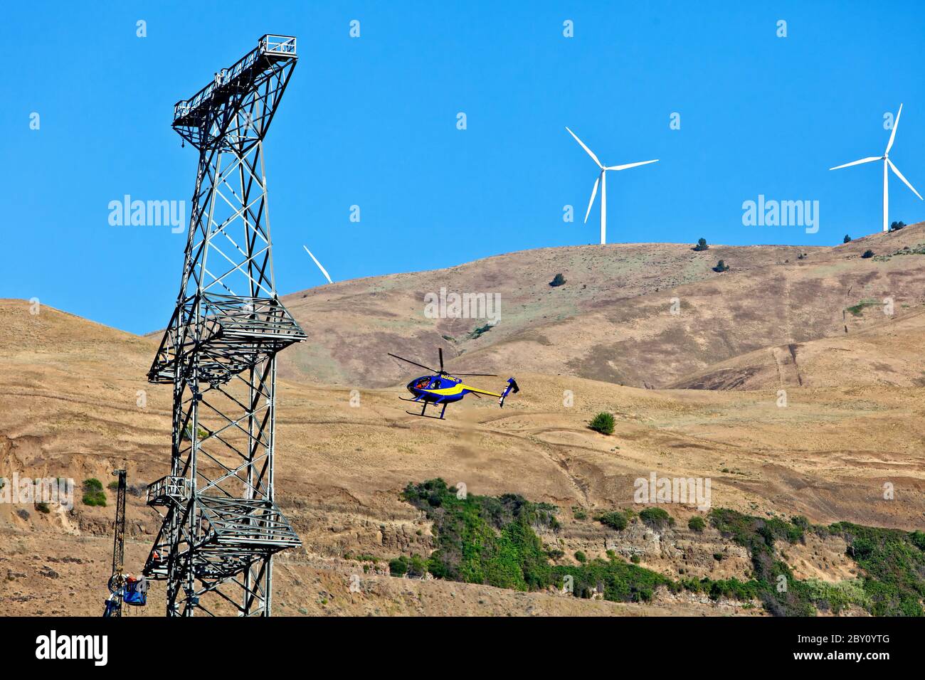 Helicopter transporting parts/material to working crew on new tower, electical transmission tower.  Hughes 500  helicopter,  Wind Farm in background. Stock Photo