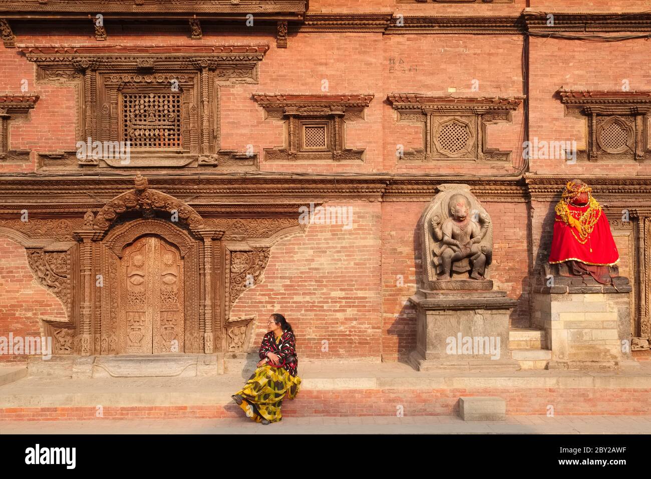A woman sits in front of the royal palace, Durbar Square, Patan (Lalitpur), Nepal, near a red-clothed Hanuman statue (r) & a frieze of Bhairav (Shiva) Stock Photo