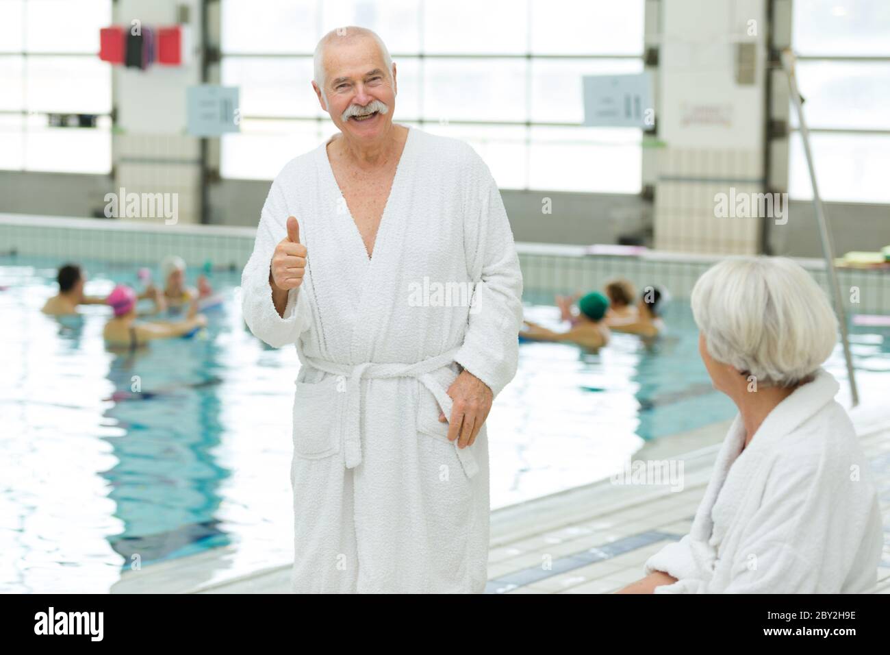 elderly couple by pool wearing bathrobes man with thumbs up Stock Photo