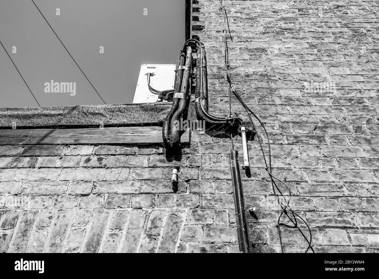 Vertical, monochrome view of current and old pipes including an air conditioning unit seen atop of a flat roof behind on old brick-built building. Stock Photo