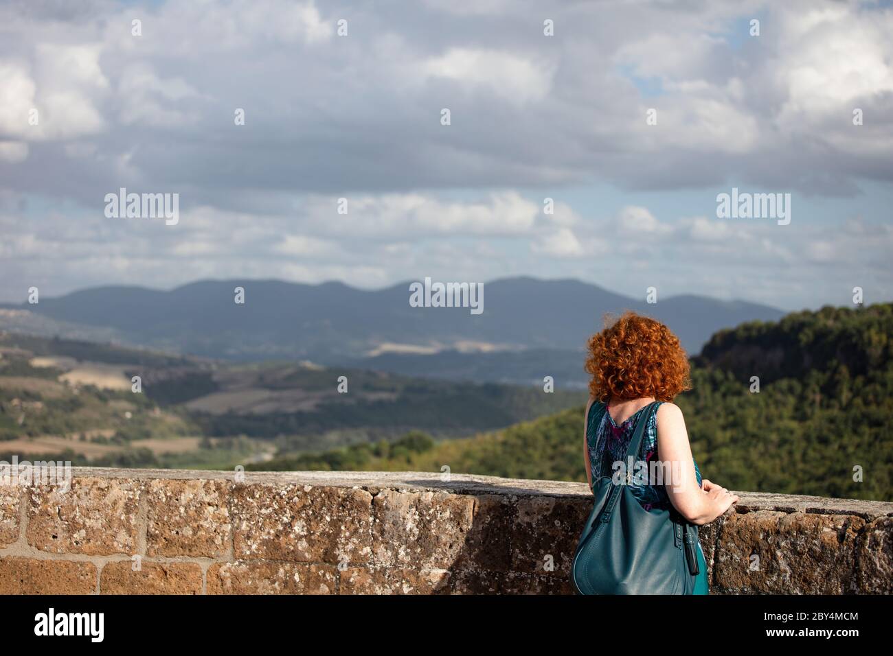Woman with curly ginger hair standing looking over a stone wall at an Italian landscape in Orvieto, Umbria. Stock Photo