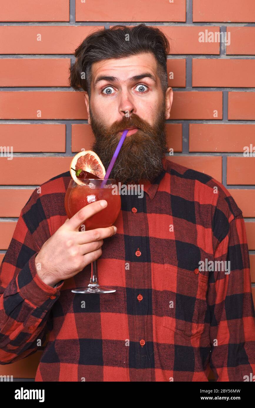 Hipster enjoys drink or cocktail. Barman with beard and surprised face drinks out of glass with drinking straw cocktail. Man in checkered shirt on brick wall background. Barman and cocktails concept Stock Photo