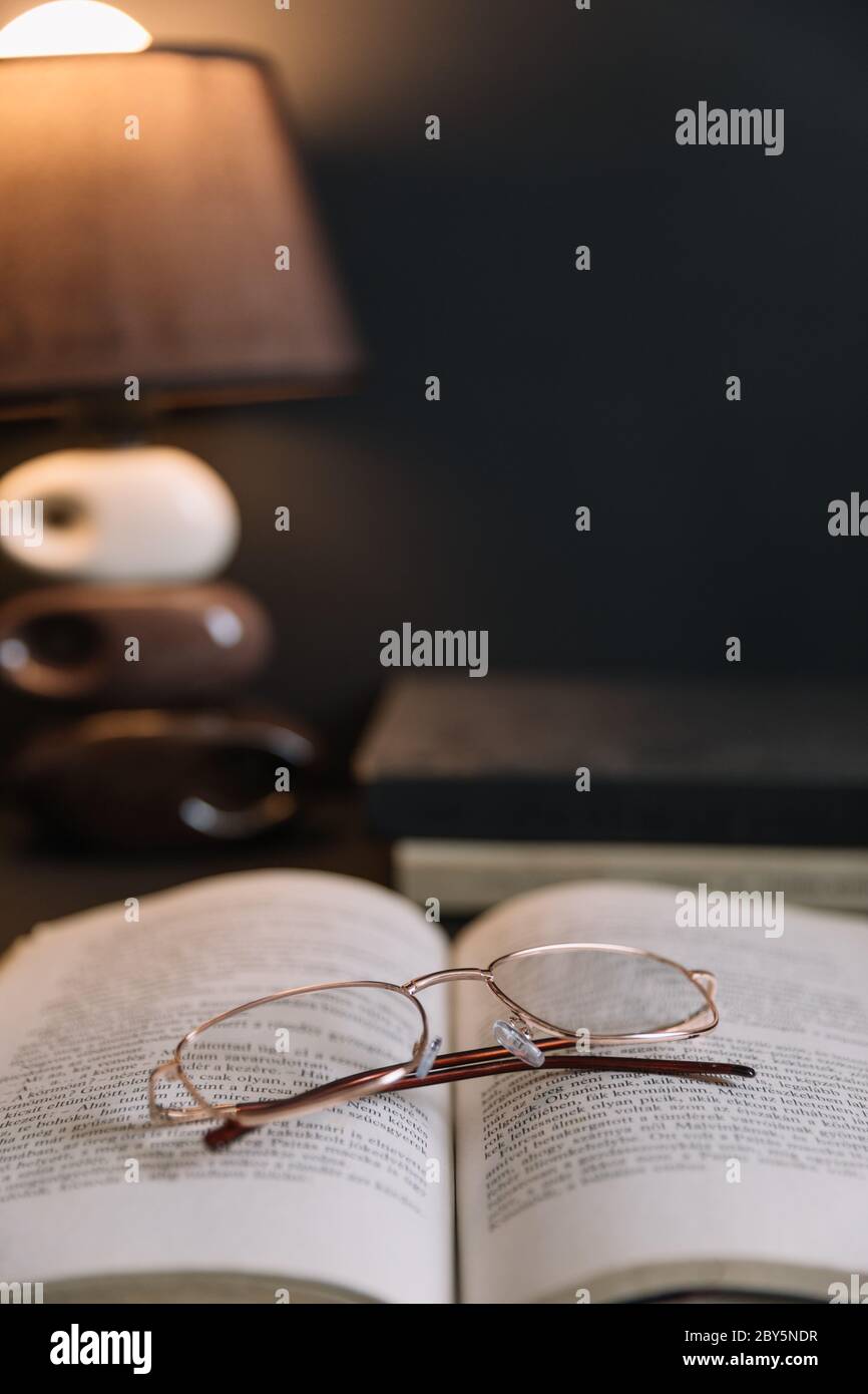 Still life with old books and reading glasses lit by night light. Calm evening atmosphere in a dark brown room. Stock Photo