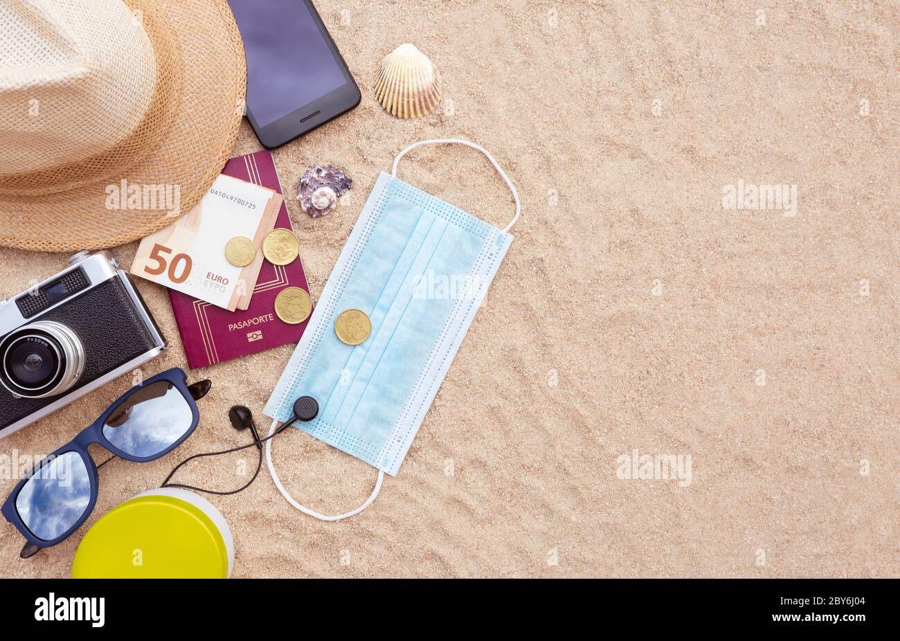 Some beach items, a protective face mask, a passport and some cash, a smartphone, hat, camera and sunglasses on the beach sand. Space for text. Concep Stock Photo