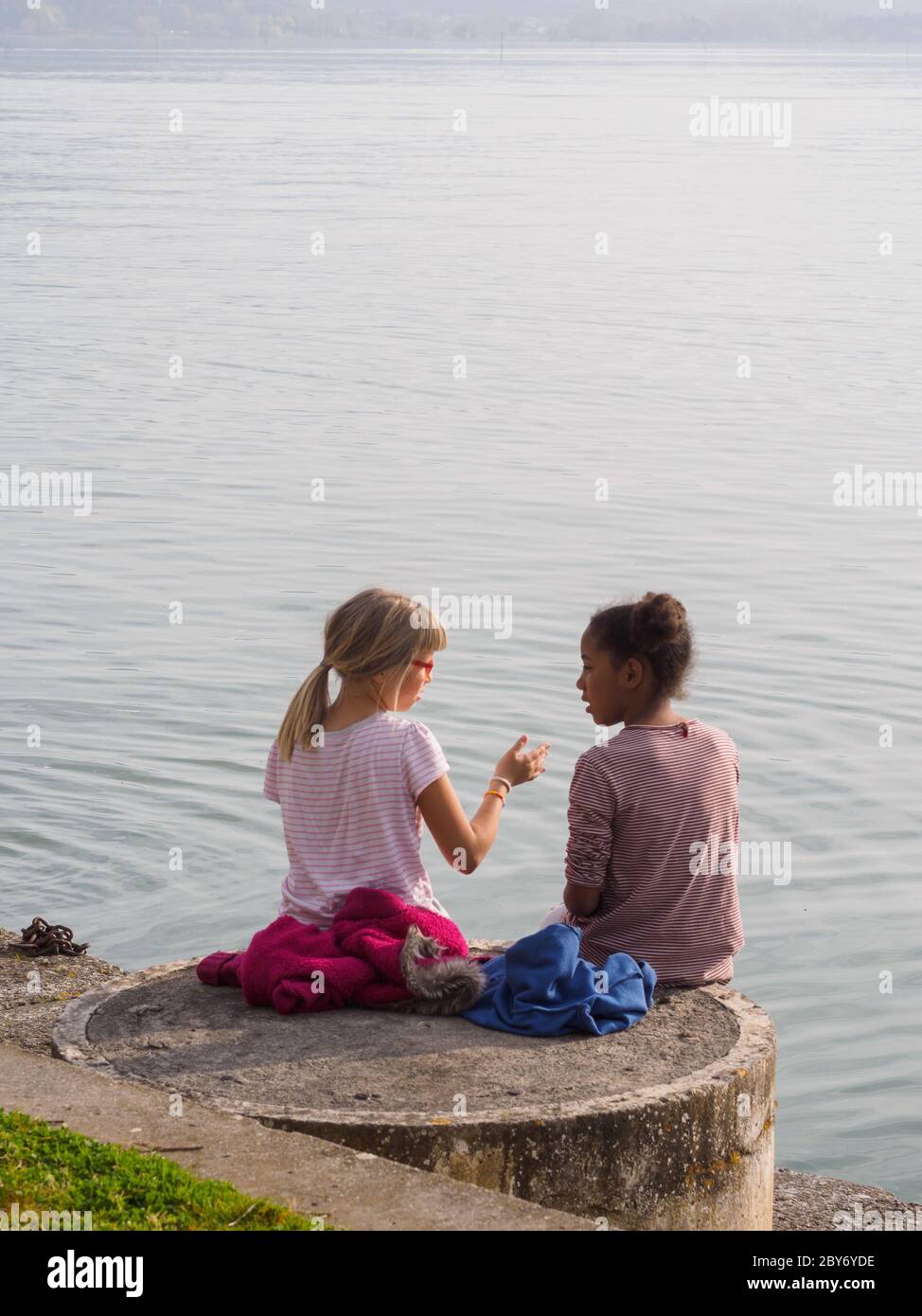 Two little girls, one blonde and the other black, sit on the edge of a lake in Italy and talk to each other... Stock Photo