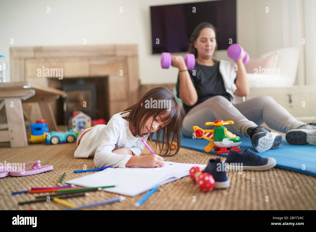 Daughter coloring while mother exercises in living room Stock Photo