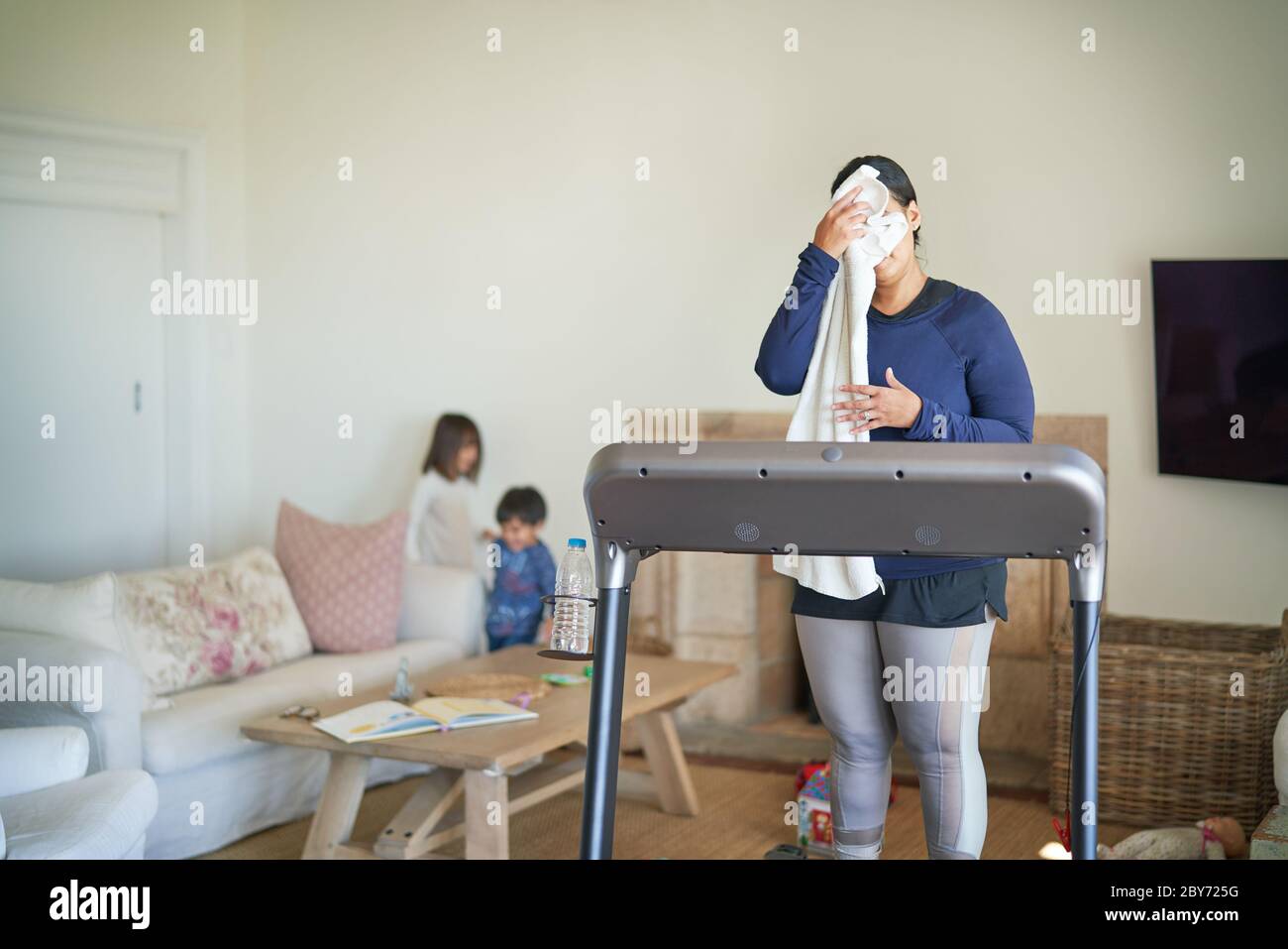 Mother on treadmill wiping sweat from face in living room with kids Stock Photo