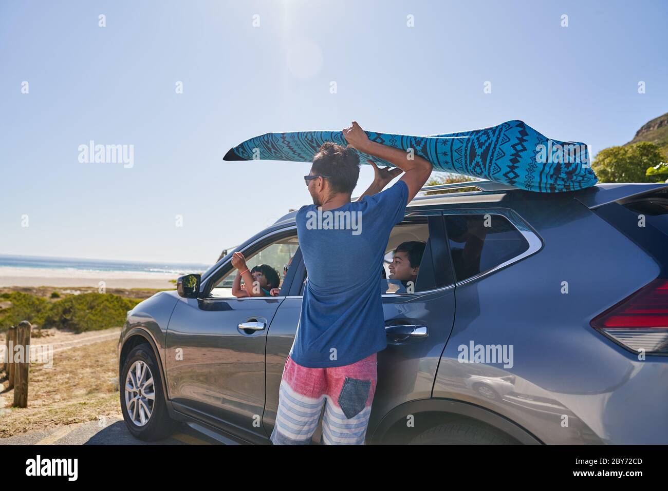 Man removing surfboard from top of car in beach parking lot Stock Photo