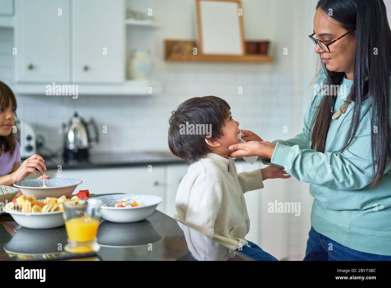 Mother wiping face of son eating fruit in kitchen Stock Photo