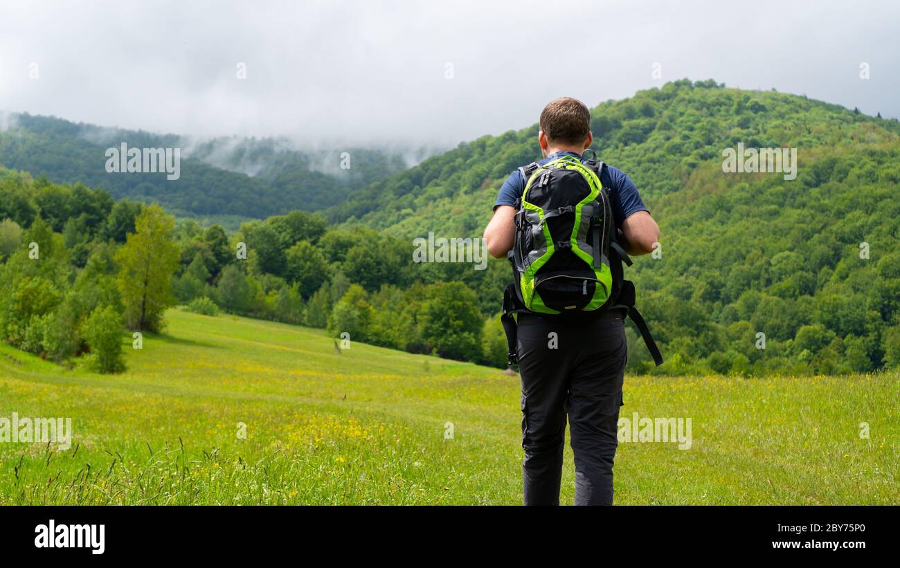 Man with green backpack hiking. Meadow overlooking the mountains and fog. Low wooded mountains. Summer weekend.  Stock Photo