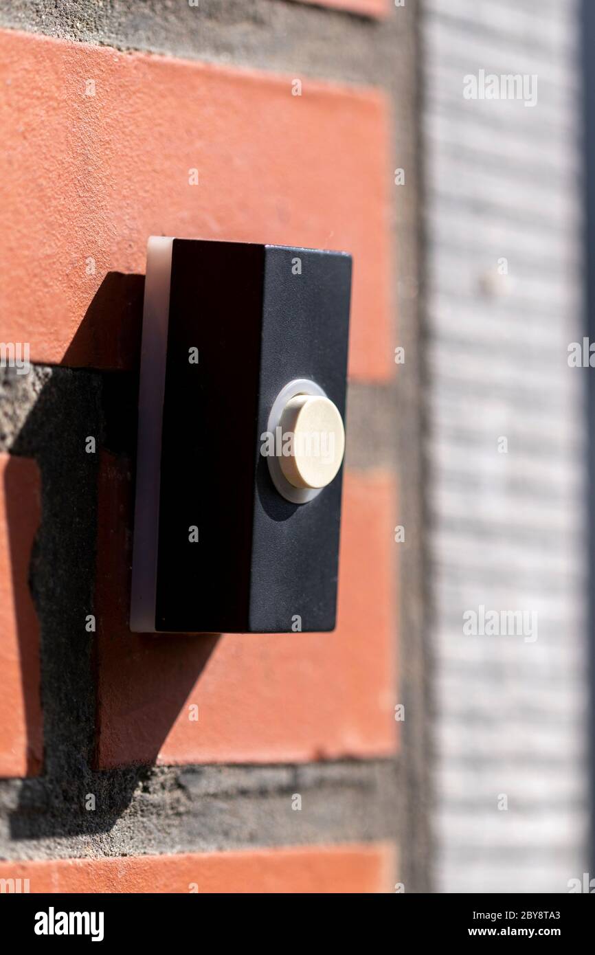 A close up portrait of a doorbell on a red brick wall. The device is next to a door and is black with a white button to be pressed to ring the doorbel Stock Photo