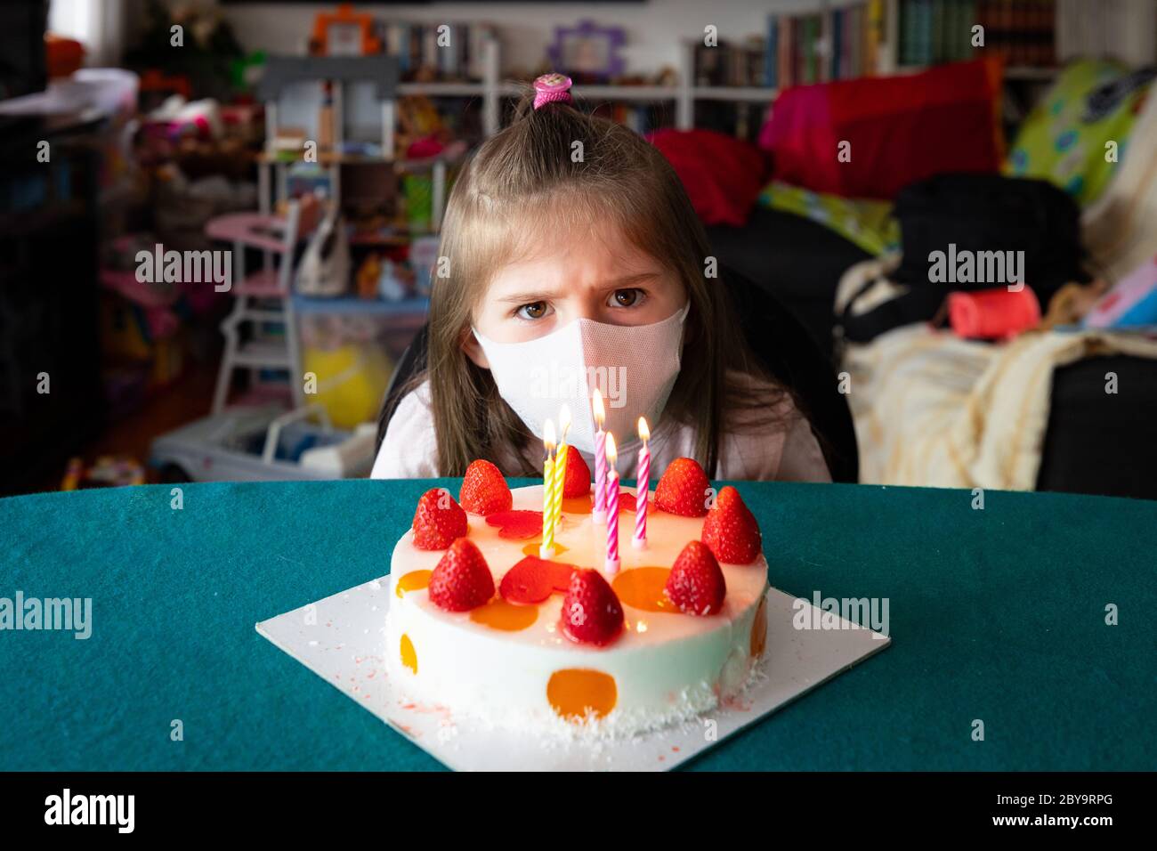 Young girl wearing a face mask during the Covid-19 or coronavirus pandemic on her birthday sitting at a table frowning as she tries to work out how to Stock Photo