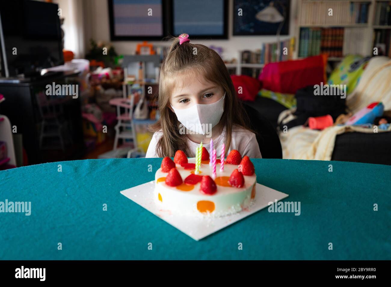 Pensive little five year old girl celebrating her birthday seated in a protective face mask at table looking quietly at her cake topped with strawberr Stock Photo
