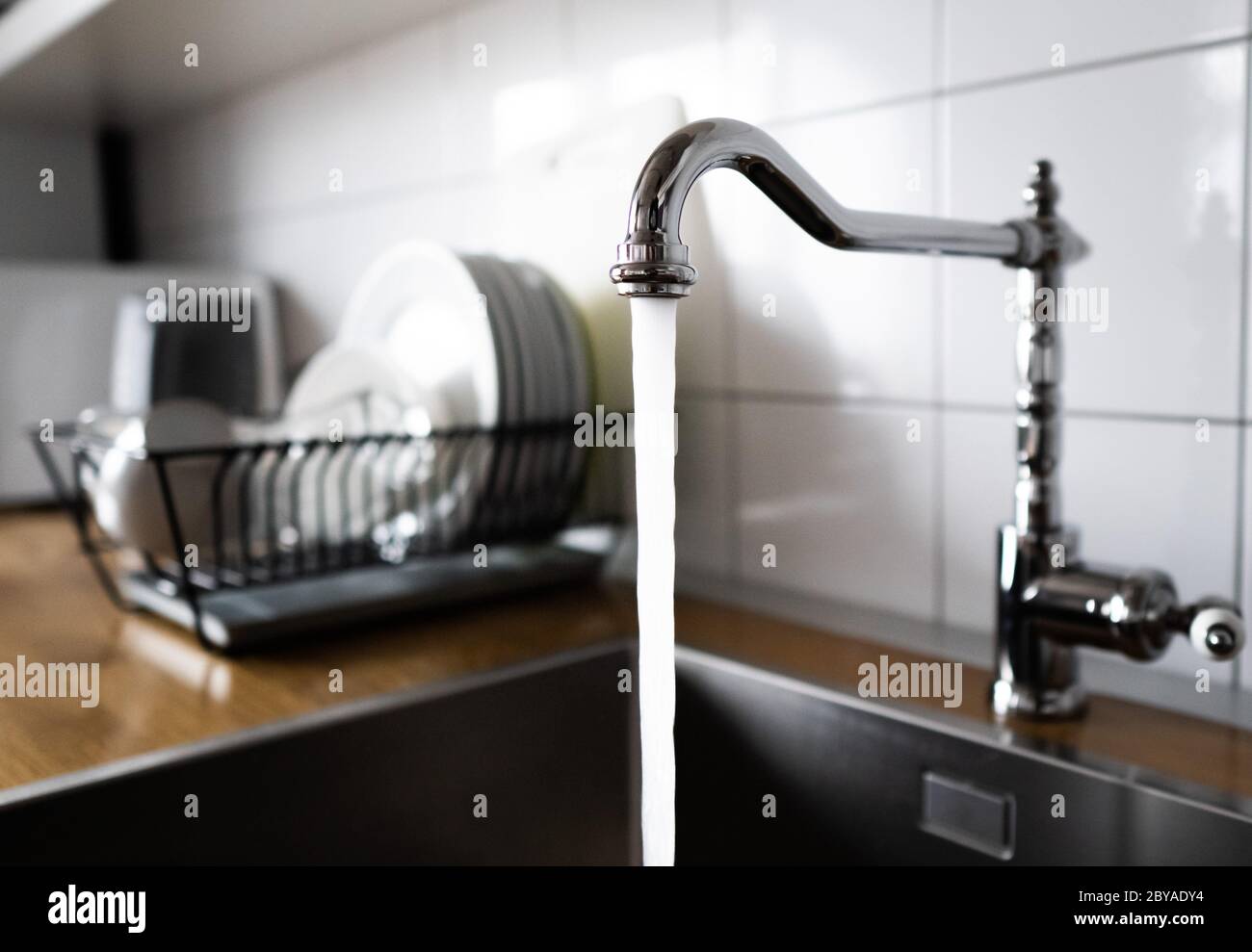 Water flowing out of a kitchen stainless steel tap into the sink. Wasting water by leaving a chrome faucet tap running. Water misuse in domestic Stock Photo