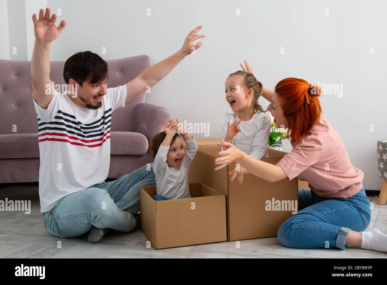 Funny active family playing on moving day, excited happy adult parents mom dad playing with cardboard boxes with cute little kids sit inside having fu Stock Photo