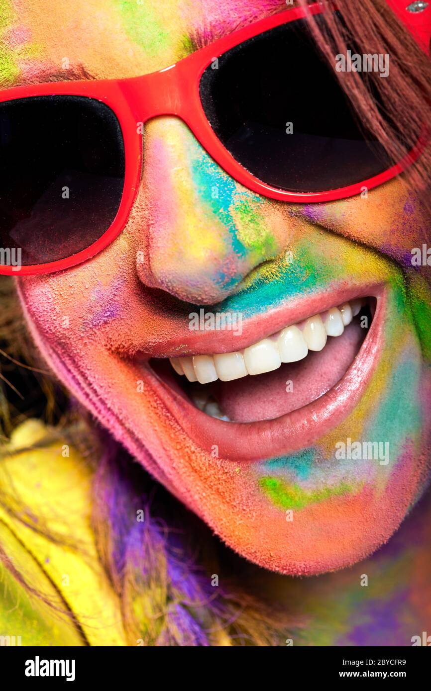 Happy excited woman face covered in rainbow colored powder. Cheerful young female in sunglasses and with face covered with multicolored bright pigment Stock Photo