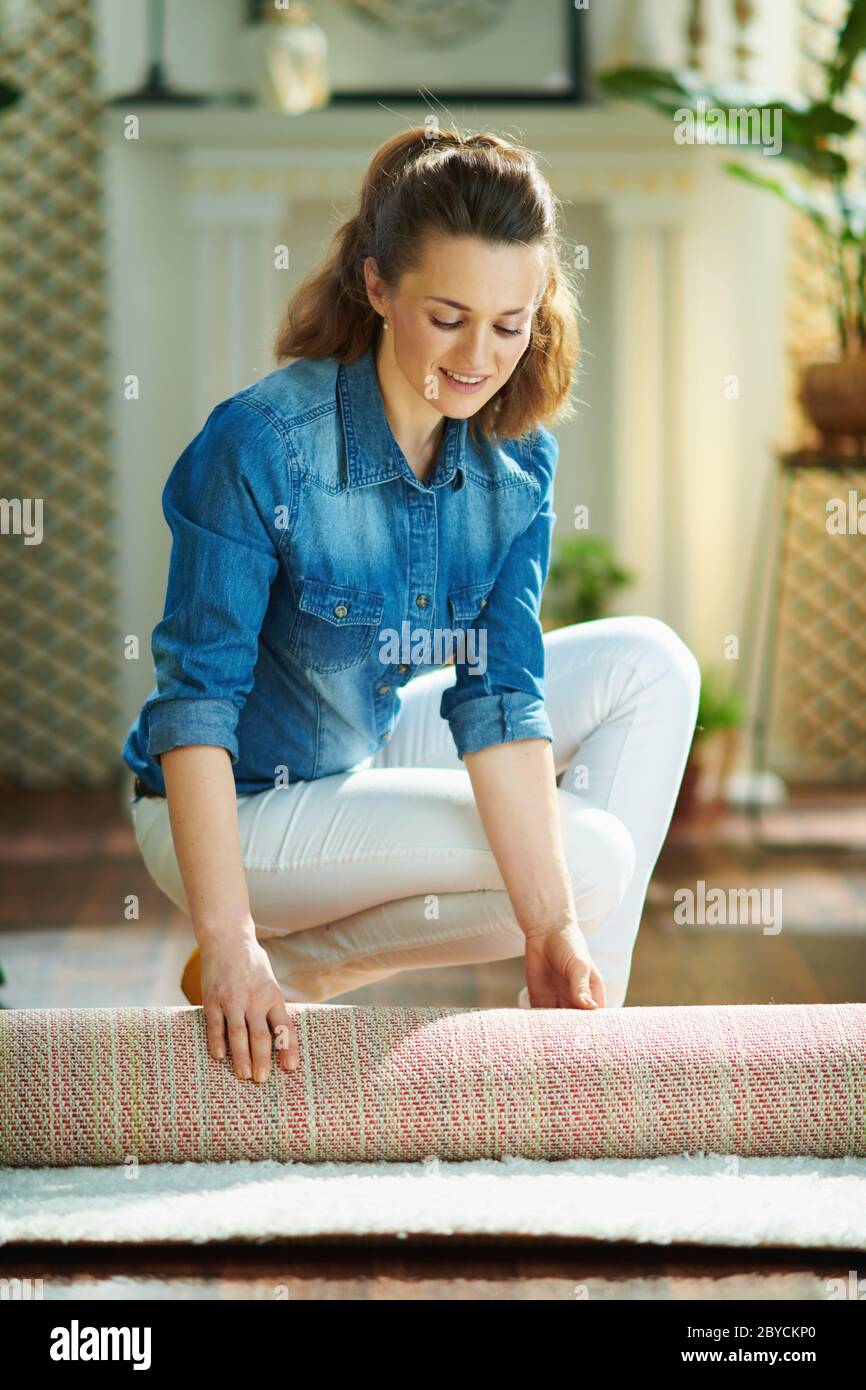 young housewife in jeans and white pants at modern home in sunny day folding carpet a roll Photo -