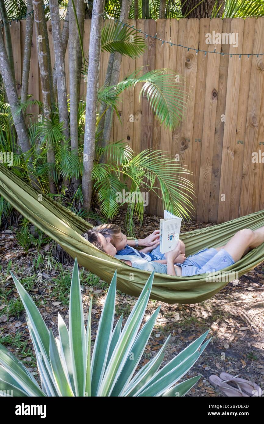 A mother and her little boy lie in a hammock and read a picture book together surrounded by greenery in their backyard in Miami, Florida. Stock Photo