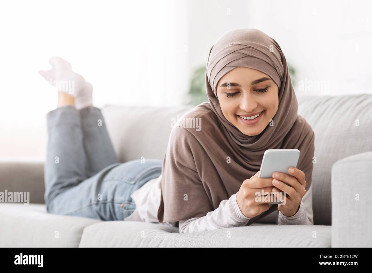 Beautiful arabic girl in hijab lying on couch and texting on smartphone ...