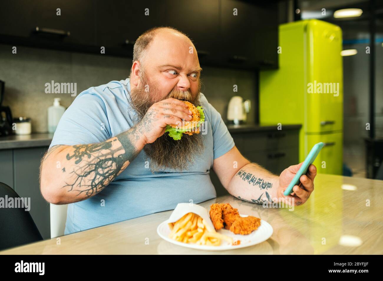 Bearded fat man looking at smartphone biting burger Stock Photo