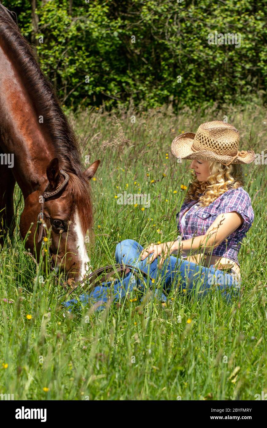 Beautiful blonde cowgirl sitting in the grass with her horse. Stock Photo