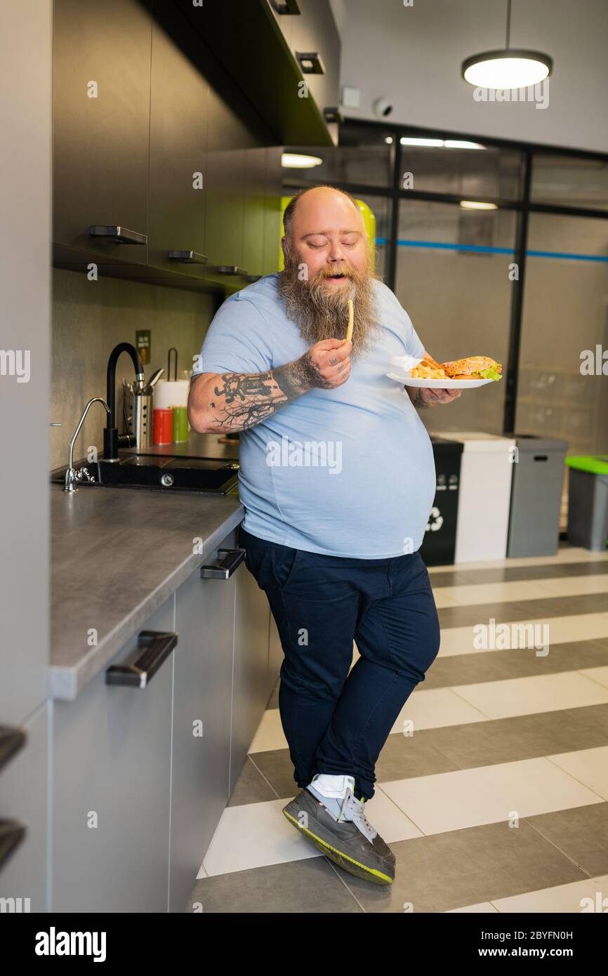 Overweight man eating french fries in the kitchen Stock Photo