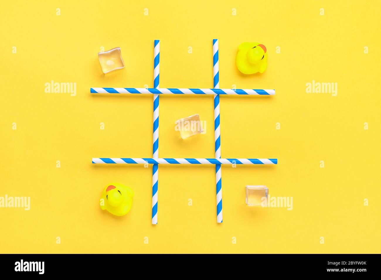 duck toy, ice cubes, paper tubes with blue stripes for drinks on a yellow background. Sea battle, tic tac toe game concept Creative composition Flat Stock Photo