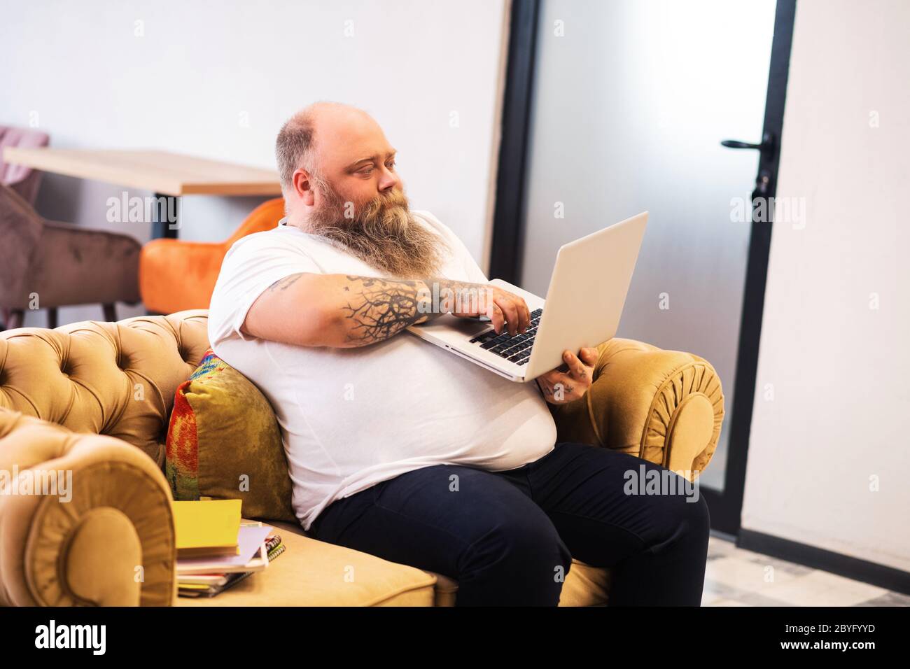 Bald bearded plump man sitting with a laptop in hands and working online Stock Photo
