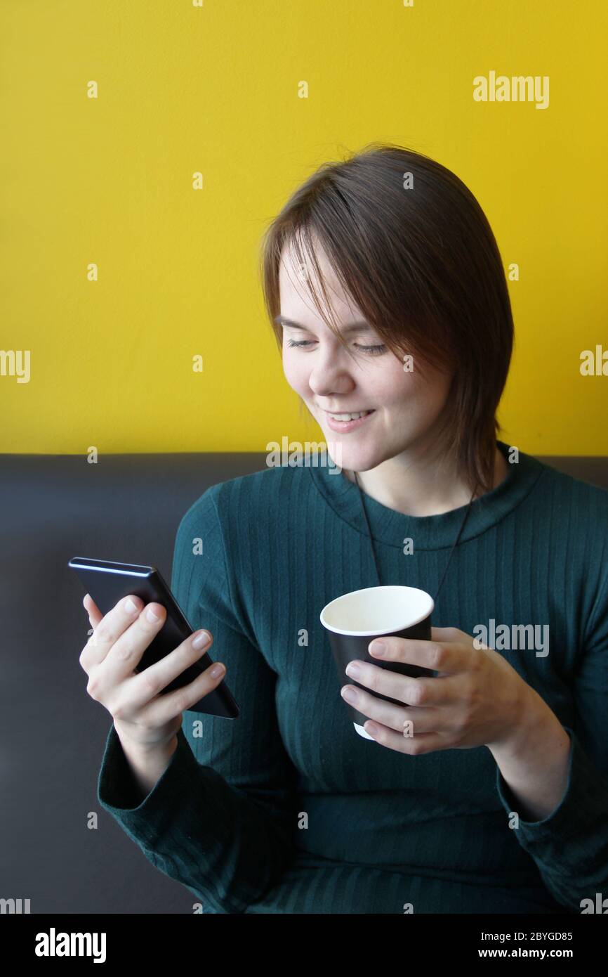 A young beautiful girl of European appearance in a blue jacket sits in a cafe with a mobile phone on the couch against the background of a yellow wall. Holding a paper cup with coffee. Stock Photo