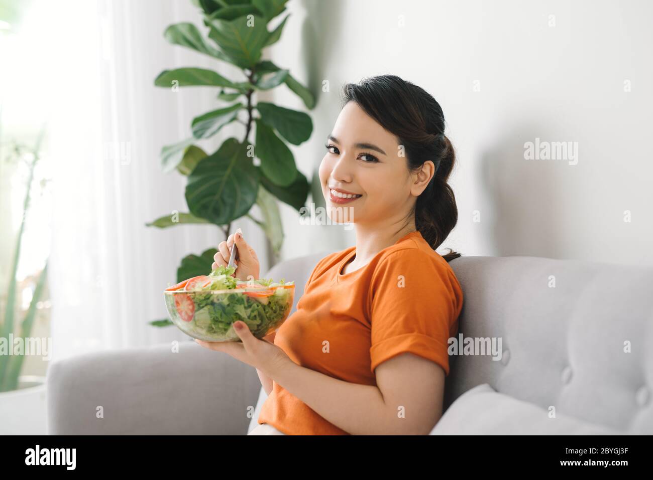 Young woman relaxing on the couch at home and eating a fresh garden salad, healthy lifestyle and nutrition concept Stock Photo
