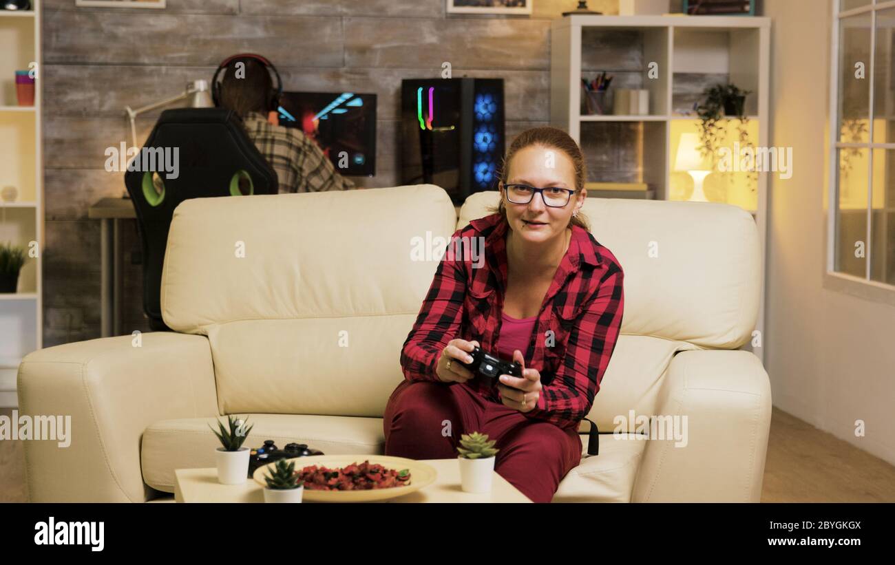 Woman sitting on couch in living room playing video games using wireless controllers. Boyfriend playing games on computer in the background. Stock Photo