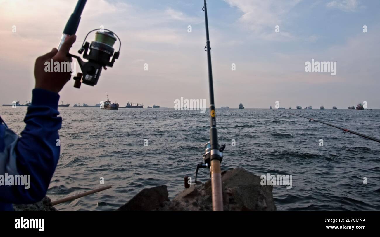 Man fishing with a spinning reel as he overlooks the Jakarta Bay from the outer wall (breakwater structure) of Port of Jakarta, Jakarta, Indonesia. Stock Photo