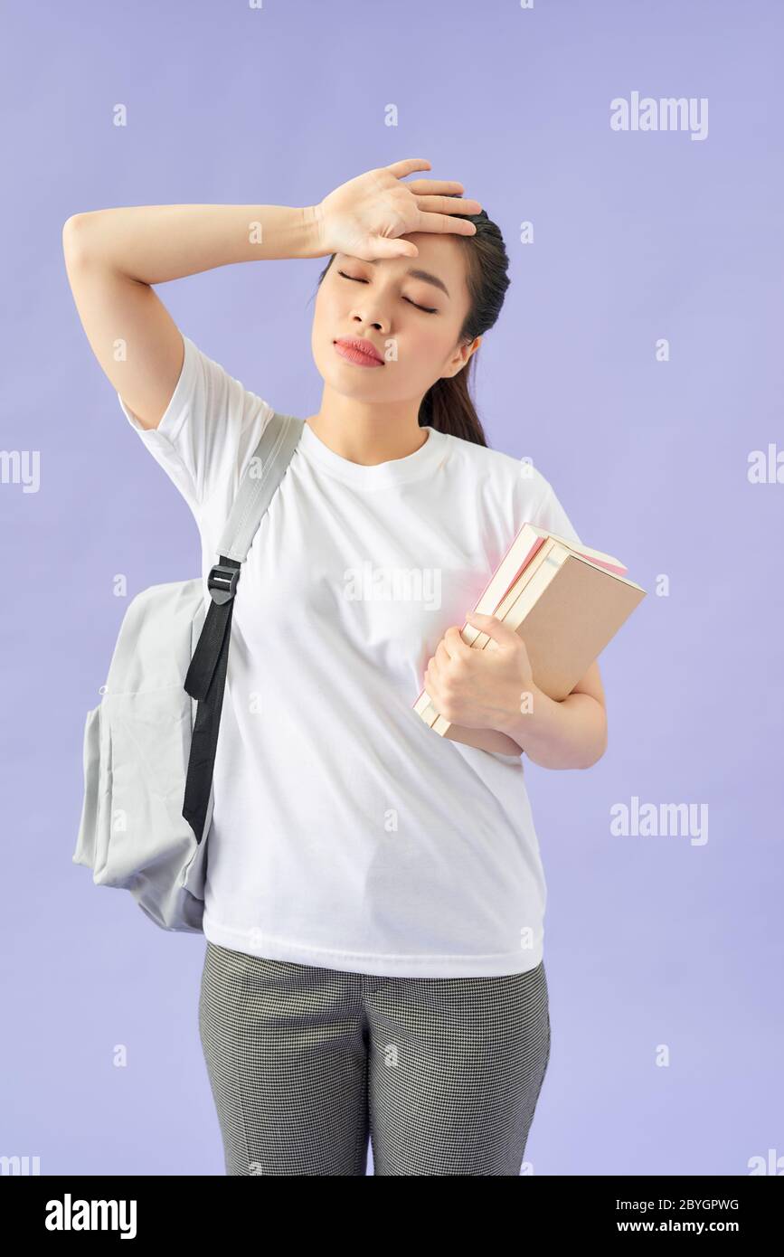 Tired young woman student with backpack on purple background.  Hold books keeping eyes closed put hand on head Stock Photo