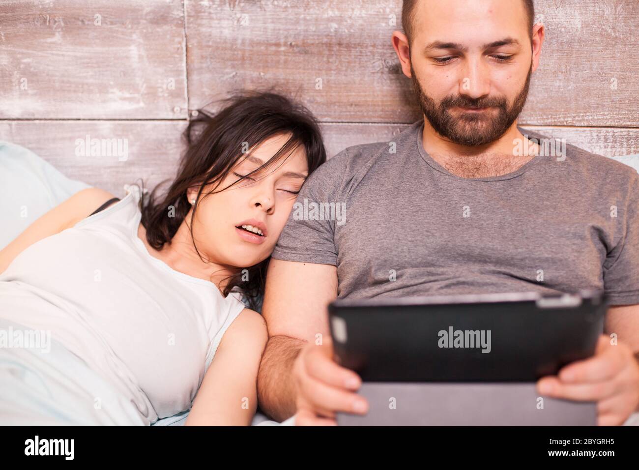 Beautiful young woman sleeping next to her husband. Man usint tablet computer. Stock Photo