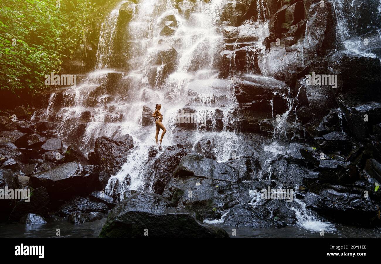 Amazing woman enjoying under stream of big and beautiful cascade waterfall. Girl with slim fit body and long black hair. Kanto Lampo in Ubud area, Stock Photo