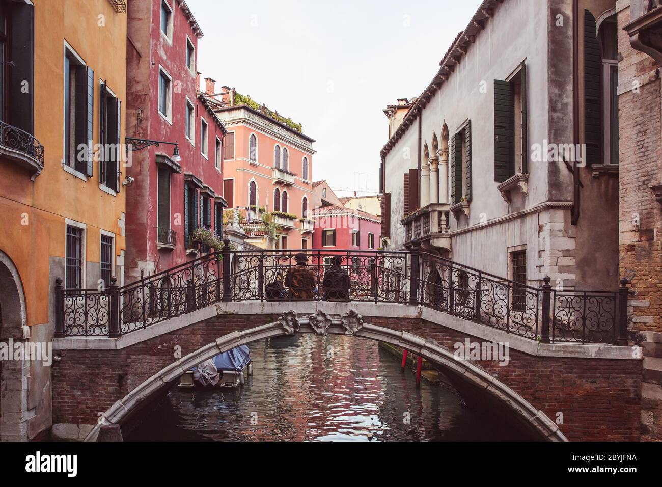 Two lovers sitting side by side on a bridge over a canal among pink buildings in Venice, Italy Stock Photo